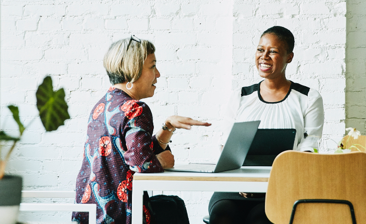 two black women smiling and talking while sitting at their laptops