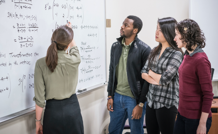 female teacher pointing to a math problem on a white board and explaining it to three adult students